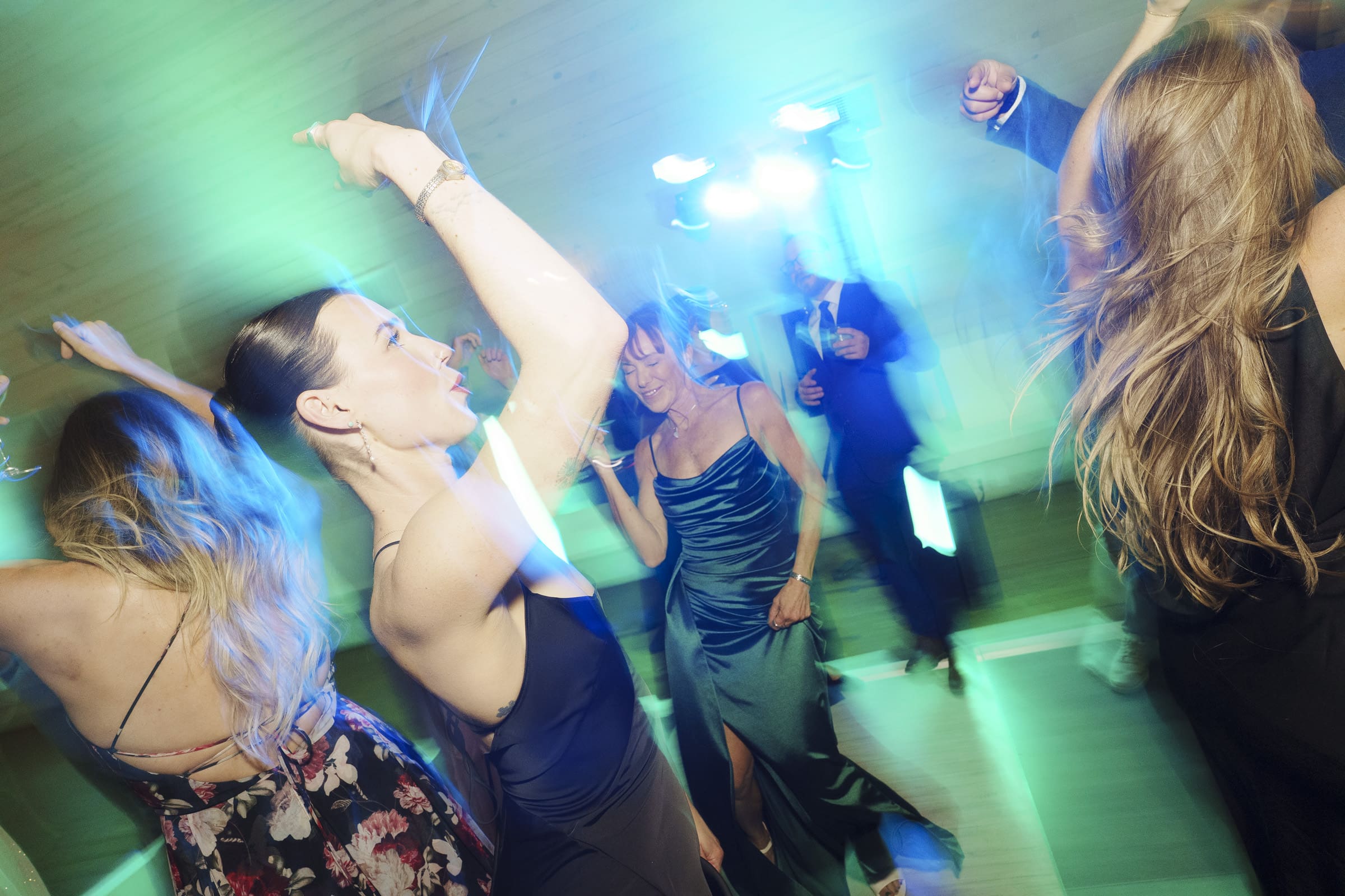 A group of people in formal attire dance energetically under bright blue and green lights at a Wakefield Barn wedding, with some raising their arms and smiling. By award-winning Raleigh wedding photographer, Joe Payne.