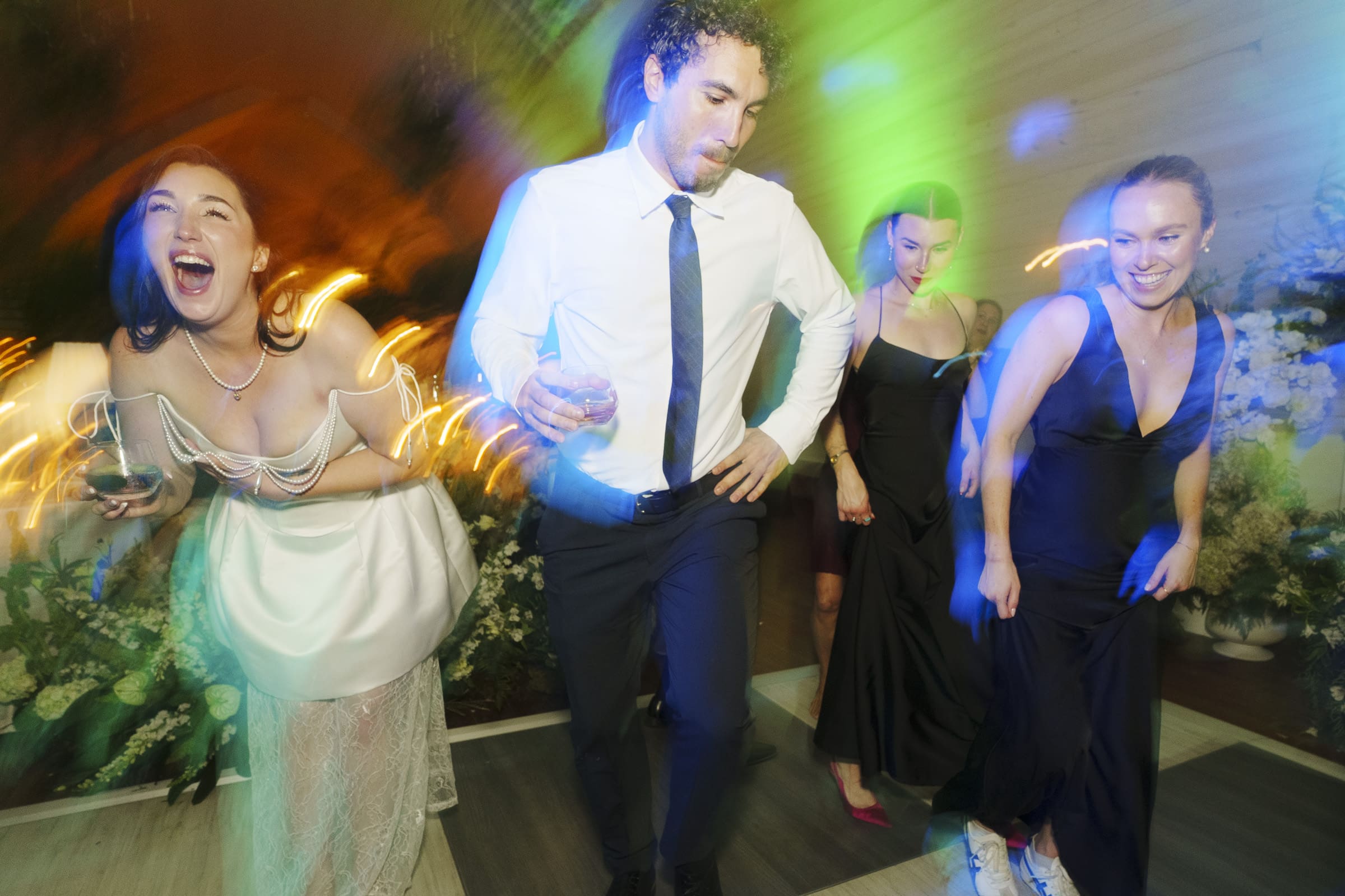 Four people in formal attire dance energetically at a barn wedding, colorful lights creating a lively, blurred effect. One woman in a white dress laughs joyfully while holding a drink; the others also appear to be enjoying themselves. By award-winning Raleigh wedding photographer, Joe Payne.