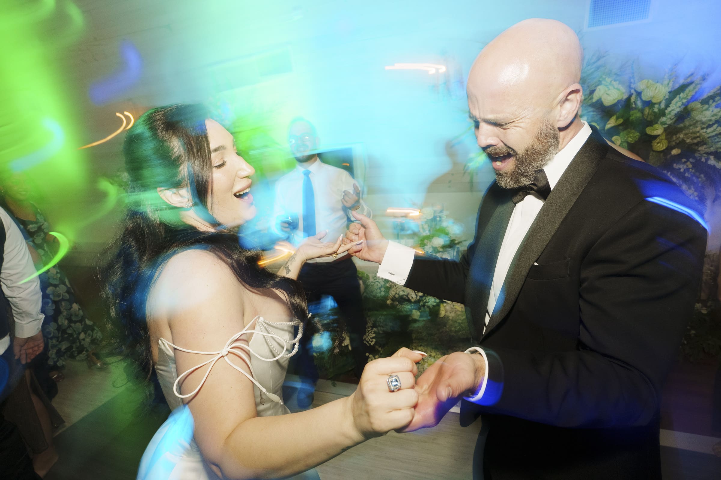 A joyful couple in formal attire dances energetically at a celebration, smiling and holding hands, surrounded by colorful, blurred lights and other guests at a lively Wakefield Barn wedding. By award-winning Raleigh wedding photographer, Joe Payne.
