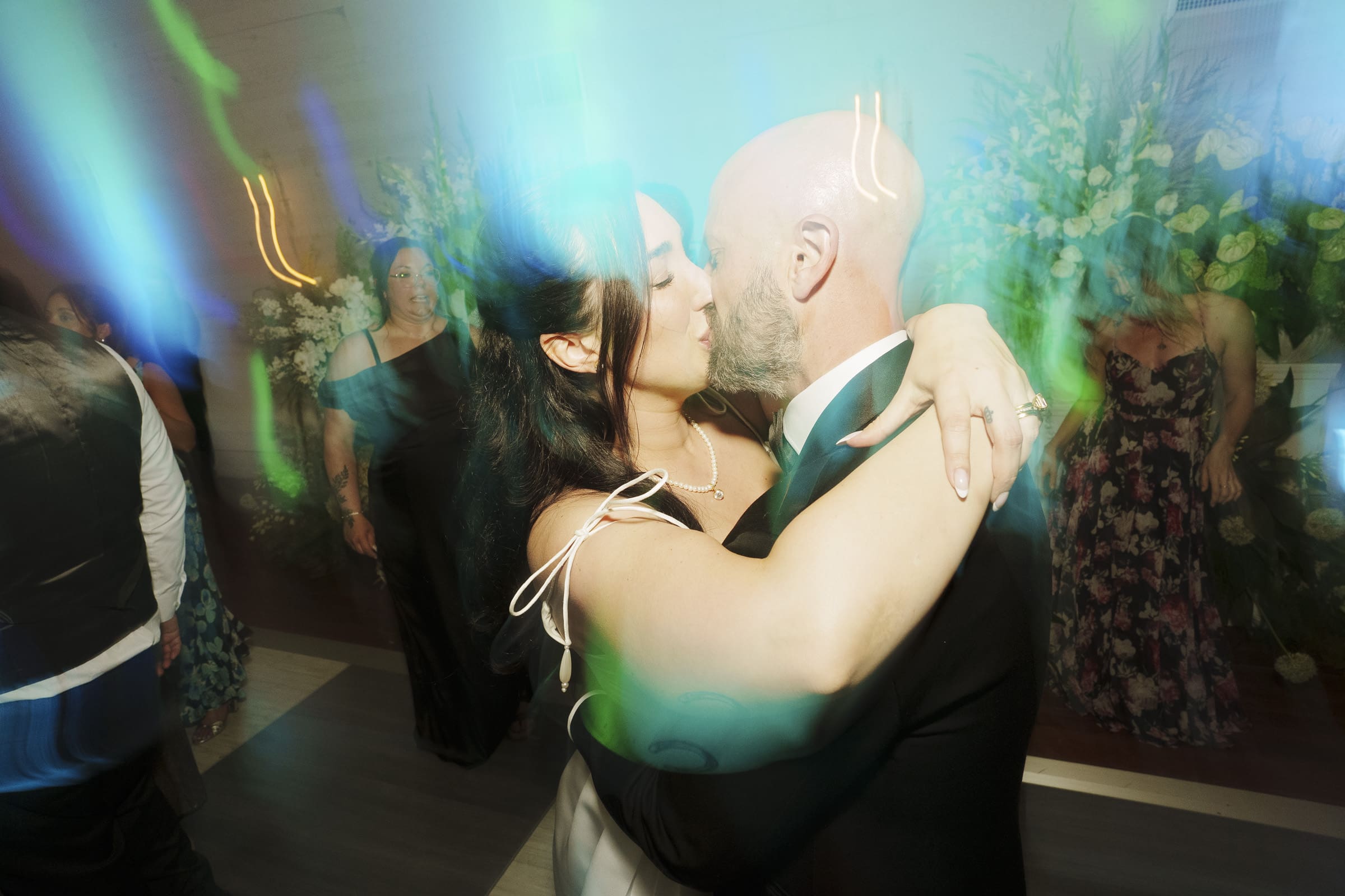 Keiko and Ryan share a kiss while dancing, surrounded by colorful light streaks and blurred guests in formal attire, creating a dreamy, celebratory atmosphere at their historic Wakefield Barn wedding. By award-winning Raleigh wedding photographer, Joe Payne.