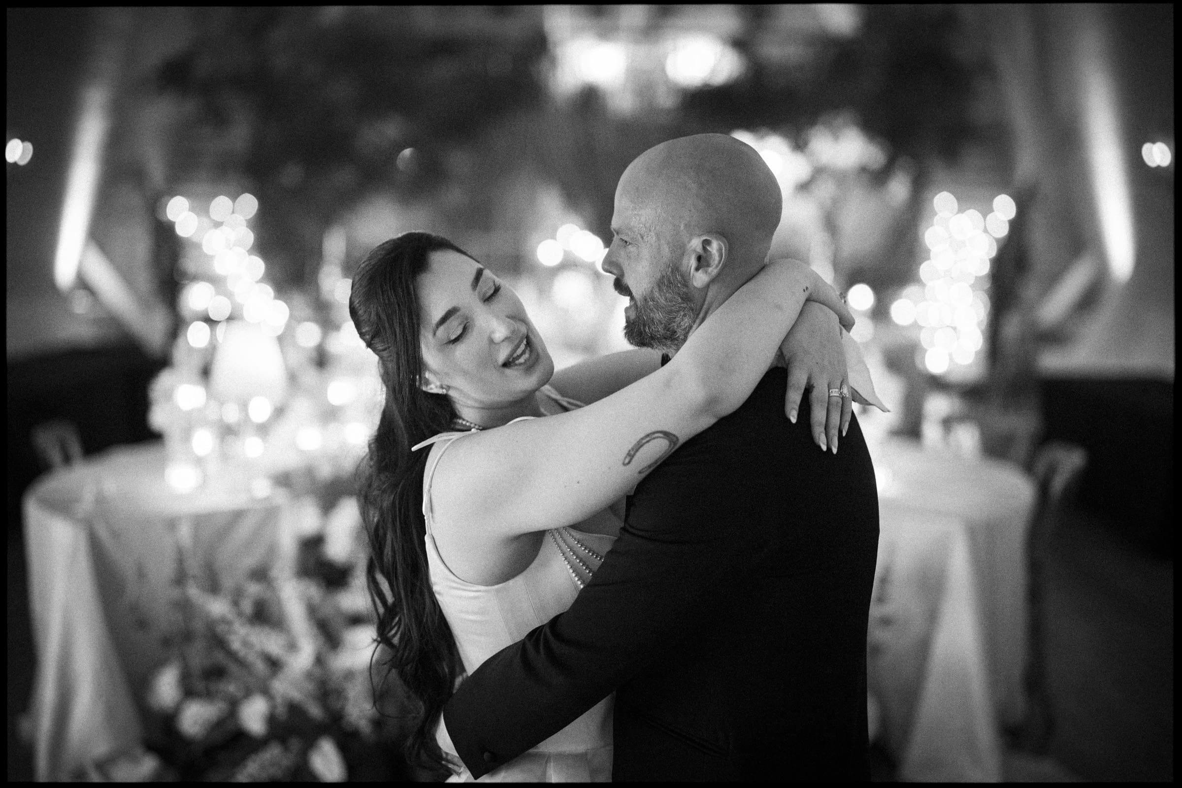 A couple embraces and dances together at a warmly lit barn wedding, possibly a reception, surrounded by round tables, candles, and soft decorative lights. By award-winning Raleigh wedding photographer, Joe Payne.