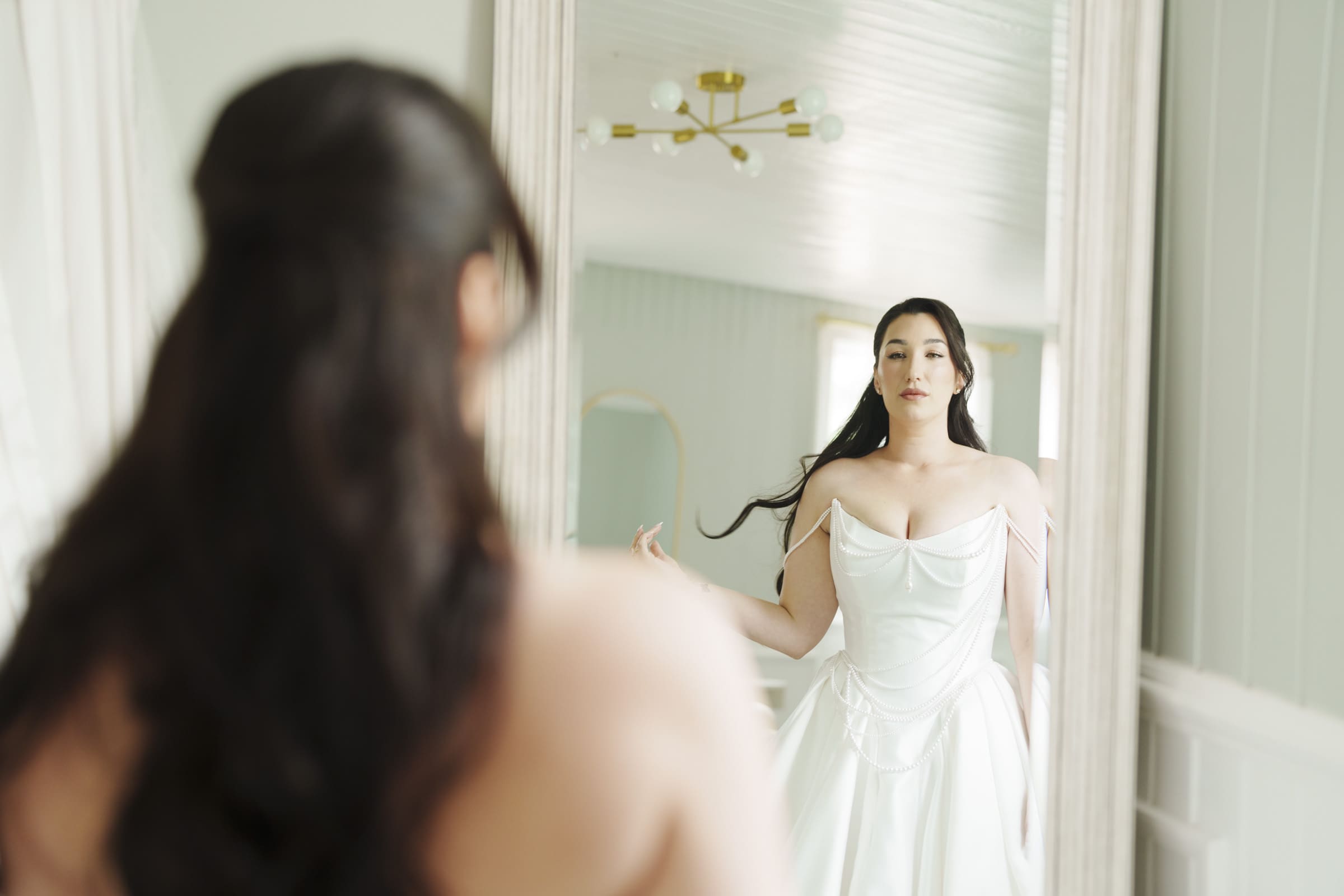 A woman in a white wedding dress gazes at her reflection in a softly lit room with pale walls and a gold ceiling light, preparing for her Wakefield barn wedding. By award-winning Raleigh wedding photographer, Joe Payne.