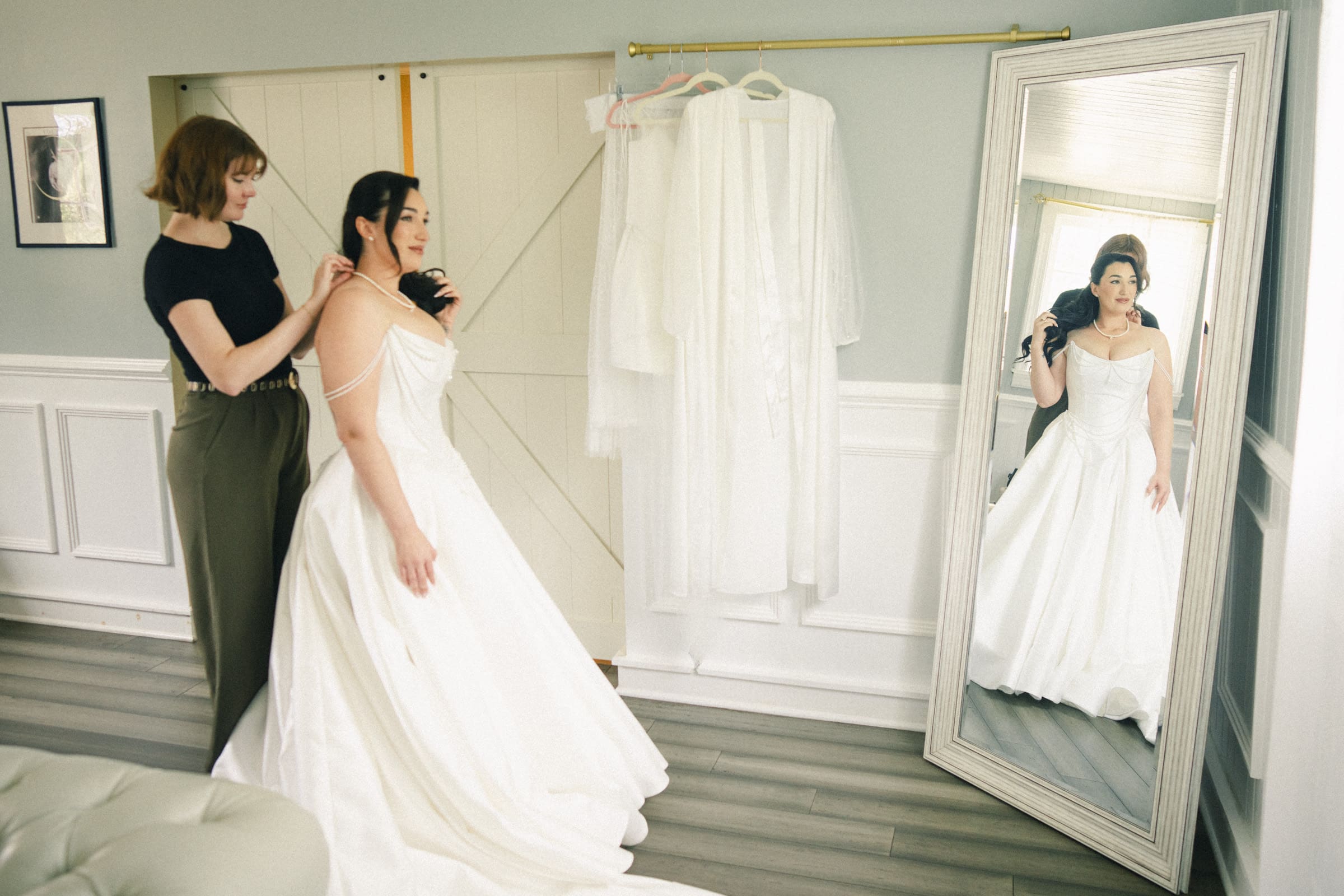 A bride in a white wedding dress stands in front of a mirror while another woman helps adjust her gown. Several white garments hang on a rod in the background of the bright, elegant room at this Wakefield Barn wedding. By award-winning Raleigh wedding photographer, Joe Payne.