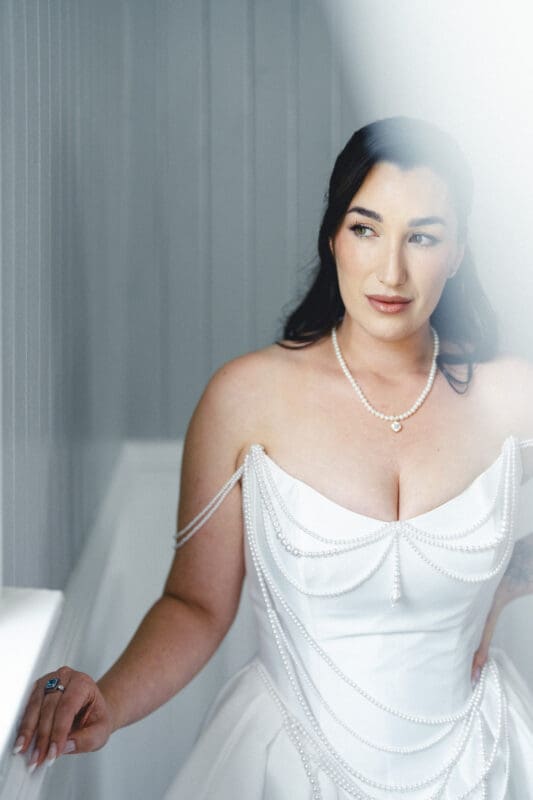 A woman in a white, off-the-shoulder wedding dress adorned with pearl strands stands indoors by a paneled wall at Wakefield Barn, looking thoughtfully to the side. Soft, natural light highlights her face and dress at this historic wedding venue. By award-winning Raleigh wedding photographer, Joe Payne.