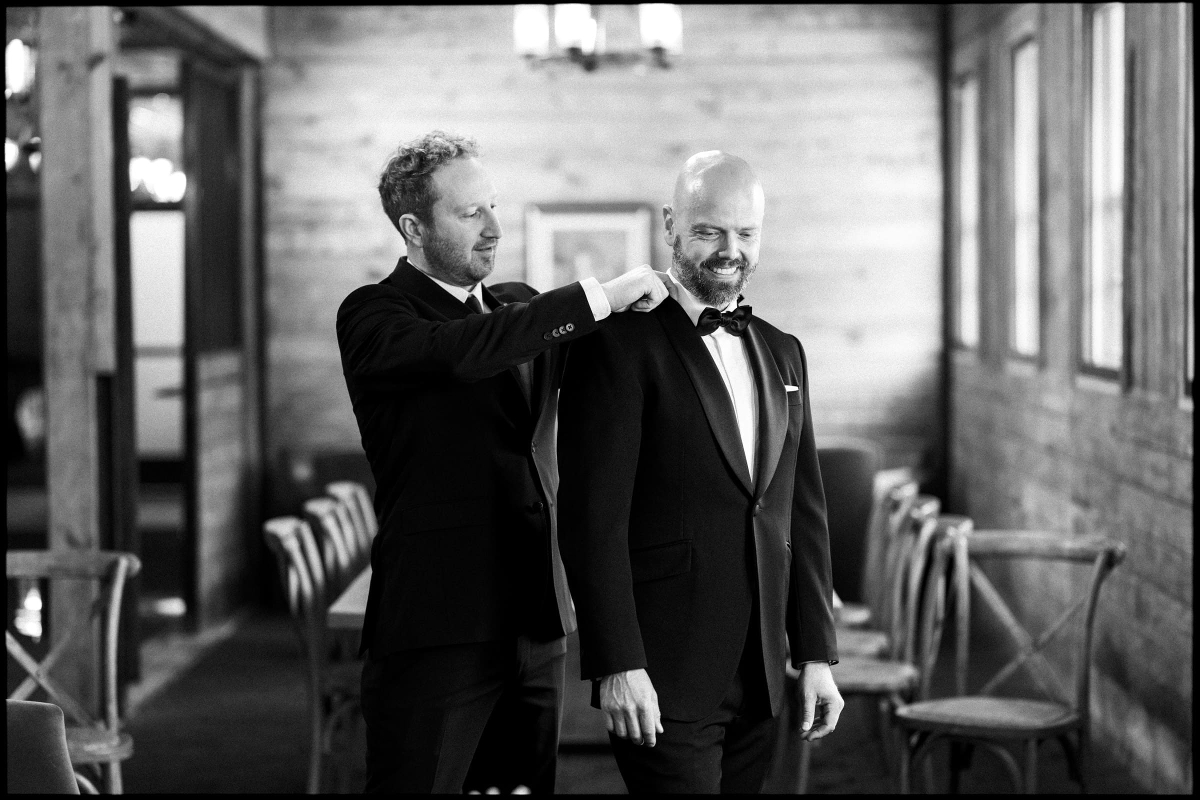 Two men in tuxedos stand in a rustic, wooden room at a Wakefield Barn wedding. One man adjusts the other’s bow tie as they smile. Empty chairs and tables are visible in the background. The photo is black and white. By award-winning Raleigh wedding photographer, Joe Payne.