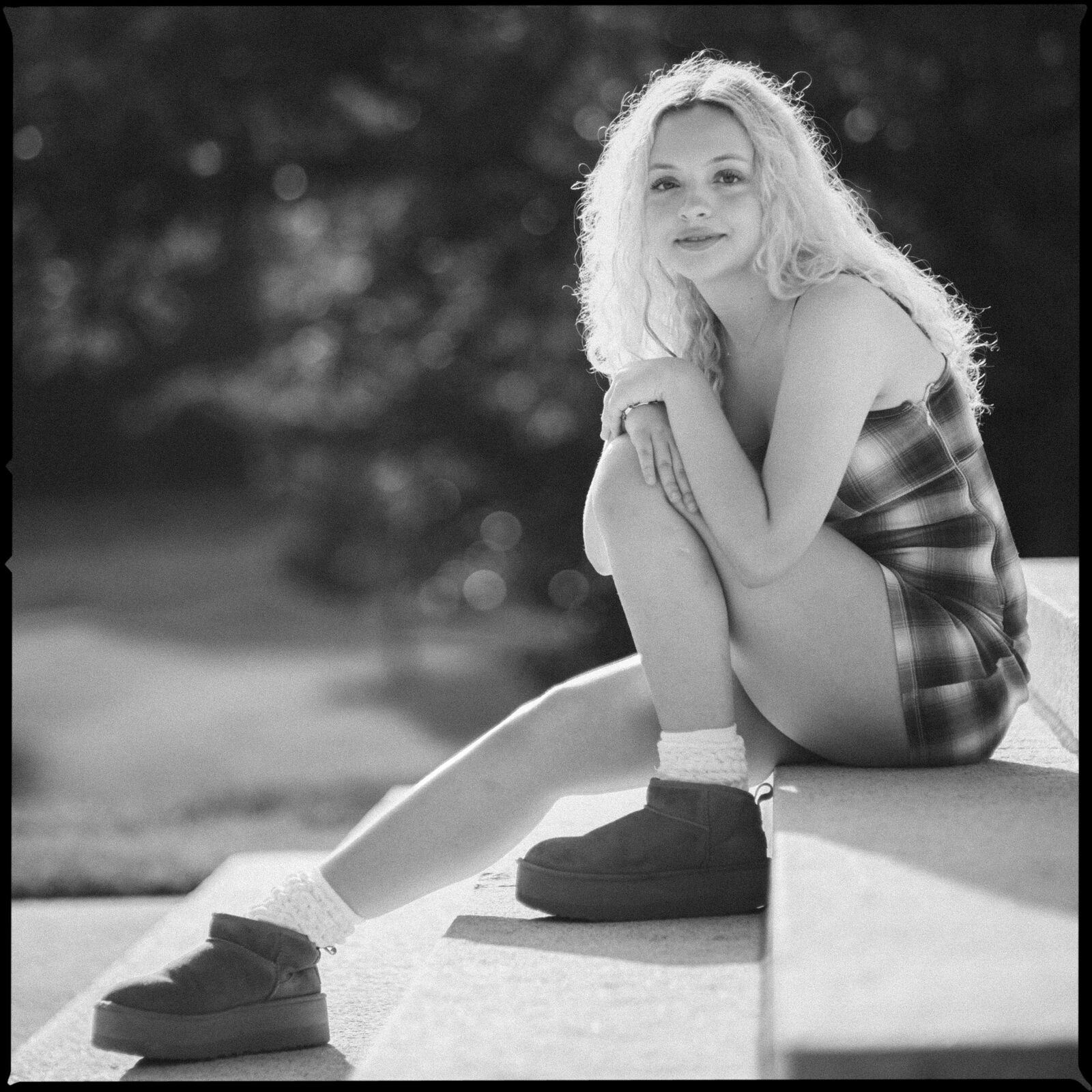 A woman with long, curly blonde hair sits on outdoor steps, wearing a plaid dress, socks, and boots. She is looking at the camera and smiling softly. The background is blurred greenery. Captured in timeless black and white by a Raleigh Durham Senior Photographer, this portrait beautifully highlights her natural charm. By award-winning Raleigh wedding photographer, Joe Payne.