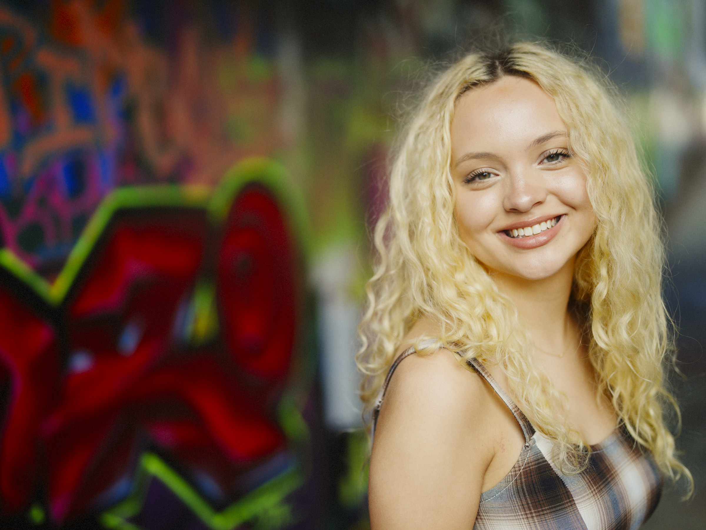 A young woman with long, curly blonde hair smiles at the camera. She is wearing a plaid tank top and stands in front of a colorful graffiti-covered wall—capturing the vibrant spirit that Raleigh senior photographers love to highlight in unforgettable senior portraits. By award-winning Raleigh wedding photographer, Joe Payne.