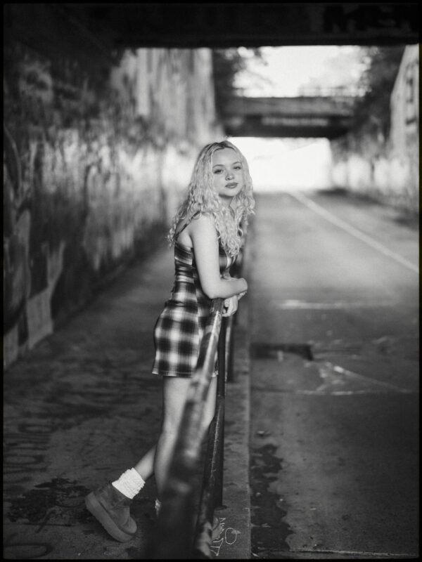 A young woman with curly hair in a plaid dress leans on a railing in an urban underpass, looking at the camera. Captured in black and white, this portrait reflects the authentic style of a Raleigh Durham Senior Photographer. By award-winning Raleigh wedding photographer, Joe Payne.