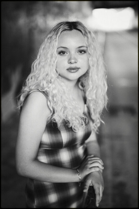 A young person with long, curly blond hair stands outdoors, wearing a plaid dress and a bracelet, with one hand resting on a railing. Captured in striking black and white, this portrait showcases the artistry of a Raleigh Durham Senior Photographer. By award-winning Raleigh wedding photographer, Joe Payne.