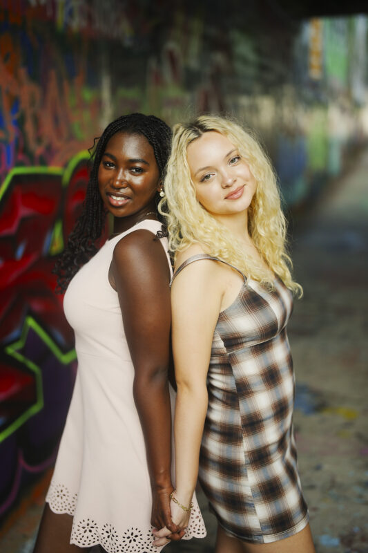 Two young women stand back-to-back, holding hands and smiling. One wears a light pink dress, the other a plaid dress. Colorful graffiti covers the wall behind them, creating a vibrant backdrop perfect for Charlotte senior photos. By award-winning Raleigh wedding photographer, Joe Payne.