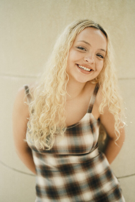Smiling young woman with long curly blonde hair, wearing a plaid sleeveless dress, stands against a light-colored wall—perfect for Charlotte Senior Photos captured by a Raleigh Durham Senior Photographer. By award-winning Raleigh wedding photographer, Joe Payne.