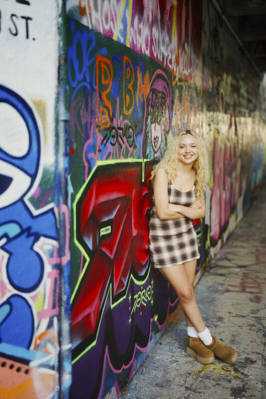 A young woman with long blonde hair in a plaid dress and boots smiles, leaning against a colorful graffiti-covered wall in an urban setting—a vibrant moment perfectly captured by a Raleigh Durham Senior Photographer. By award-winning Raleigh wedding photographer, Joe Payne.