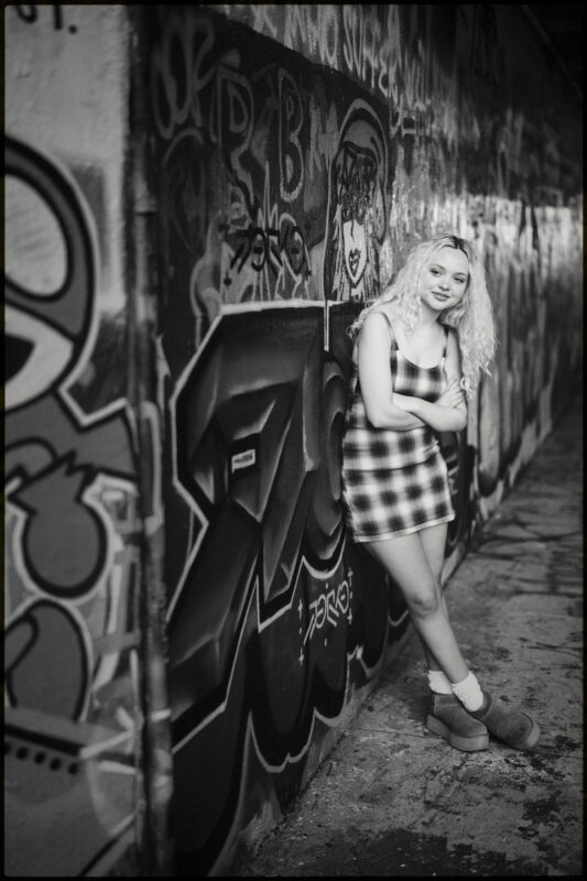 A young woman with curly blonde hair in a plaid dress leans against a graffiti-covered wall in an urban alley, smiling with arms crossed. Capturing the spirit of Charlotte senior photos, the black and white image highlights her confident presence amidst the city’s artistic backdrop. By award-winning Raleigh wedding photographer, Joe Payne.