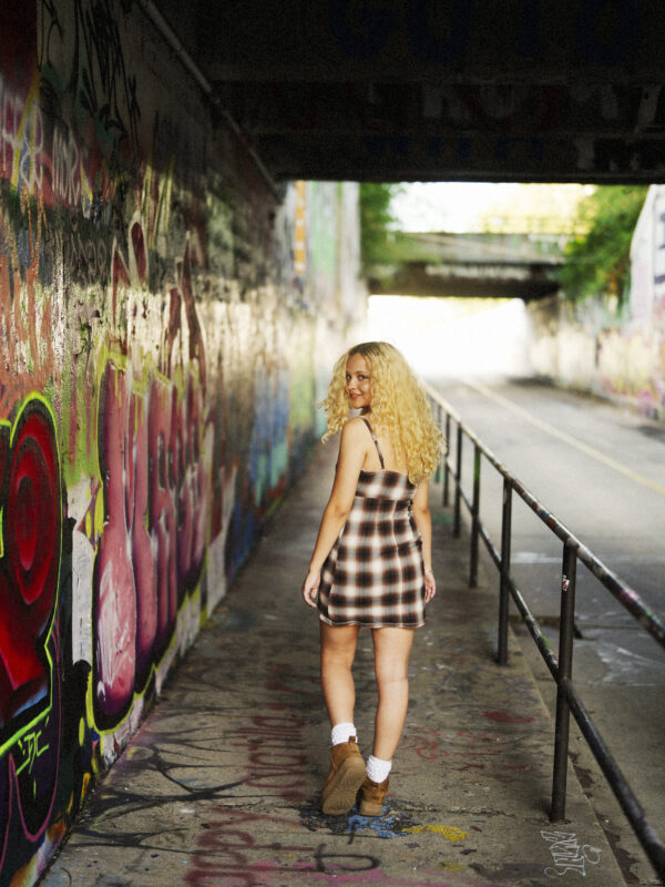 A woman with curly blonde hair in a plaid dress walks in a graffiti-covered tunnel, looking back and smiling, with sunlight ahead—an unforgettable moment captured by a Raleigh Durham Senior Photographer. By award-winning Raleigh wedding photographer, Joe Payne.
