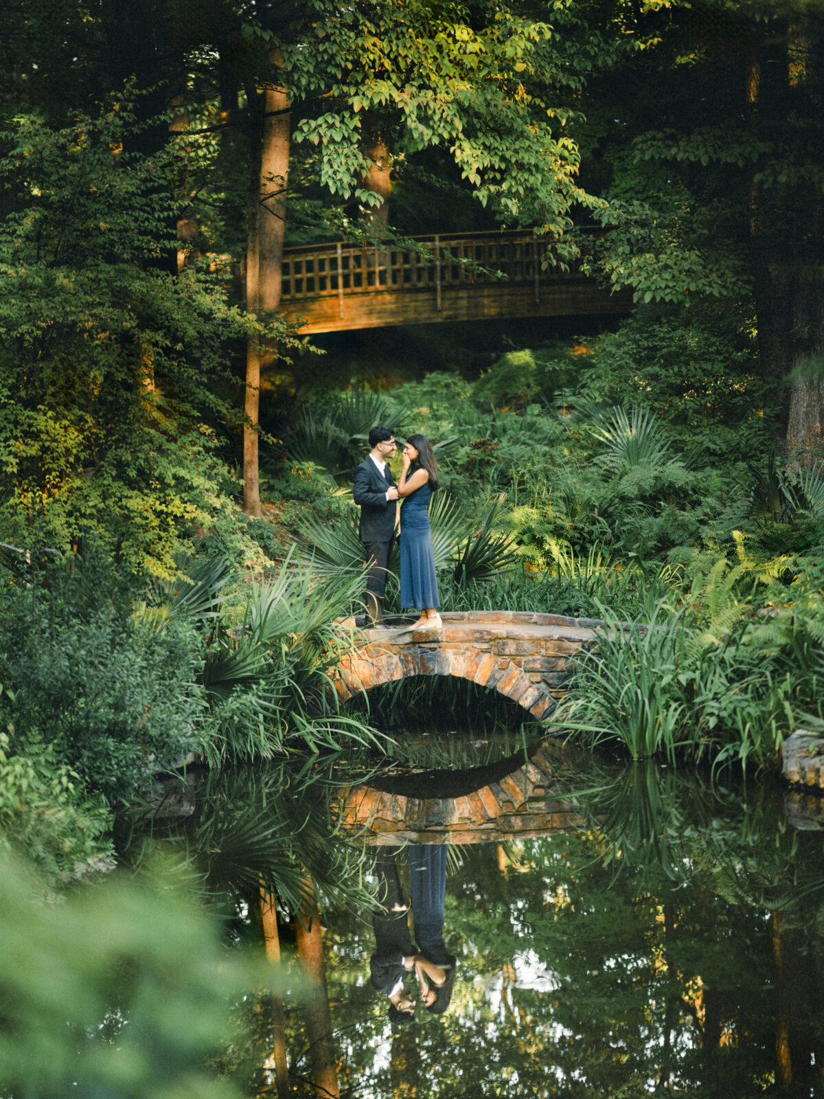 A couple stands on a small stone bridge in a lush, green garden, reflected in the water below. By award-winning Raleigh wedding photographer, Joe Payne.