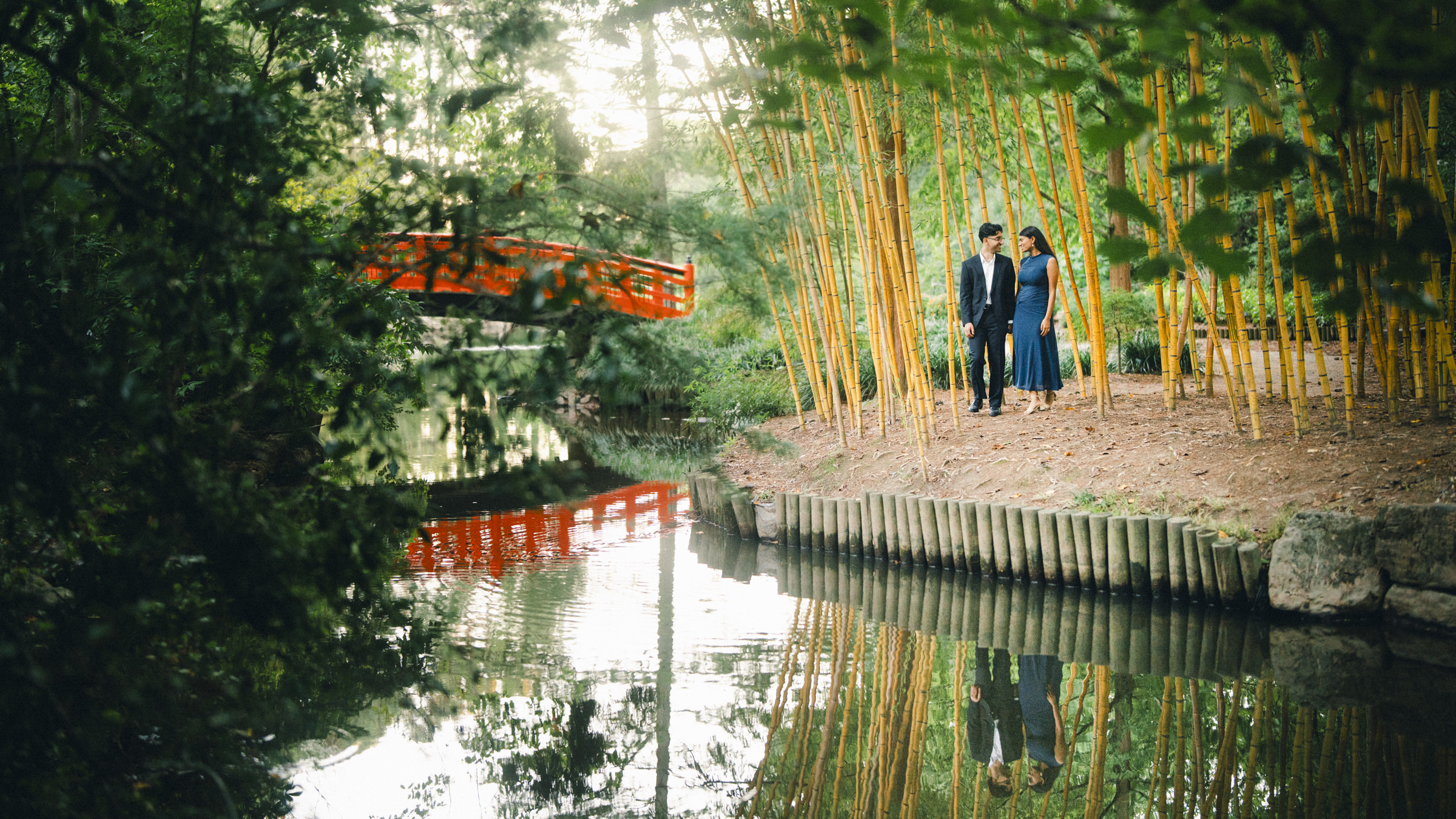 A couple stands in a Duke Gardens bamboo grove beside a pond, captured during a proposal photography session with a red wooden bridge in the background. By award-winning Raleigh wedding photographer, Joe Payne.