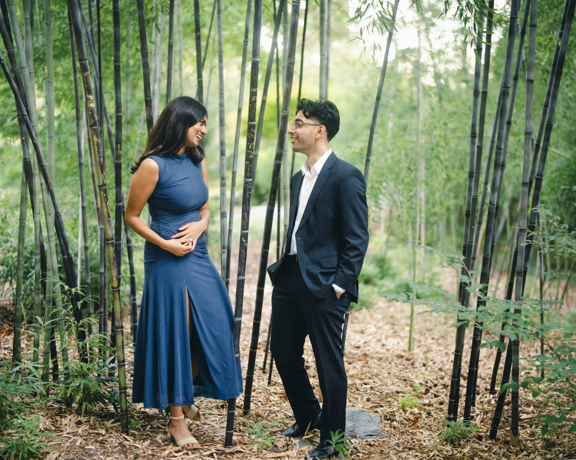 A woman in a blue dress and a man in a black suit smile at each other while standing in a bamboo grove during an engagement photography session. By award-winning Raleigh wedding photographer, Joe Payne.