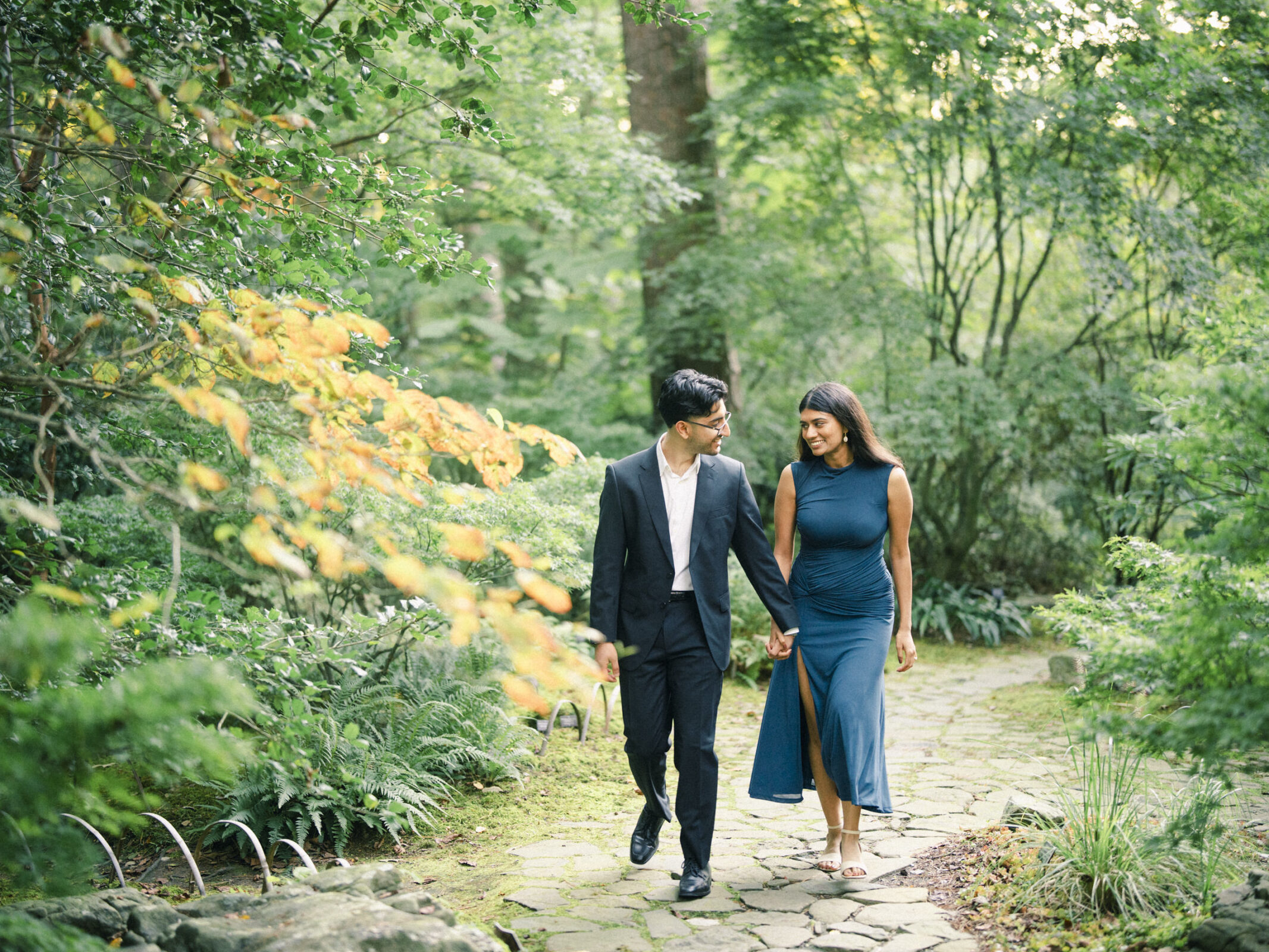 A couple in formal attire walks hand-in-hand along a stone path in a wooded Duke Gardens, captured by an engagement photographer. By award-winning Raleigh wedding photographer, Joe Payne.