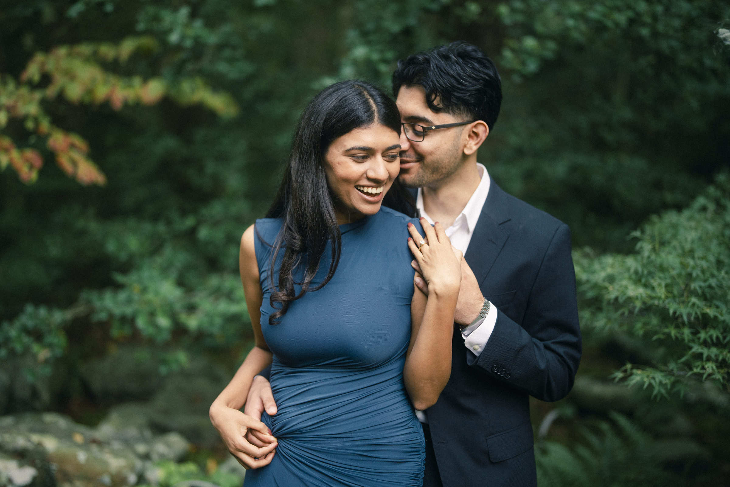 A smiling woman in a teal dress stands with a man in a dark suit who whispers in her ear during their Duke Gardens engagement photography session. By award-winning Raleigh wedding photographer, Joe Payne.