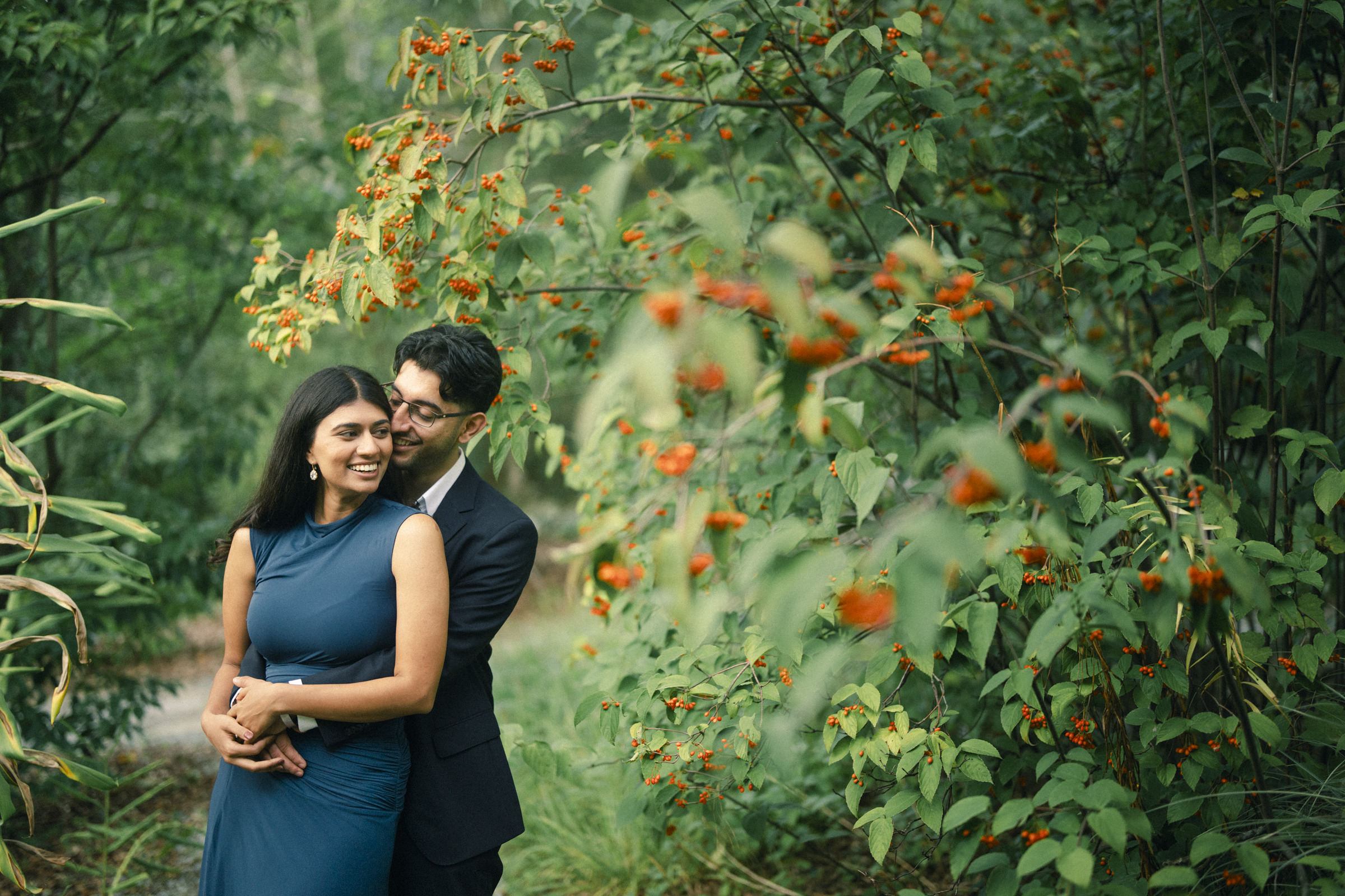 A man in a suit embraces a woman in a blue dress during an engagement photography session as they stand together in front of a shrub with orange berries. By award-winning Raleigh wedding photographer, Joe Payne.
