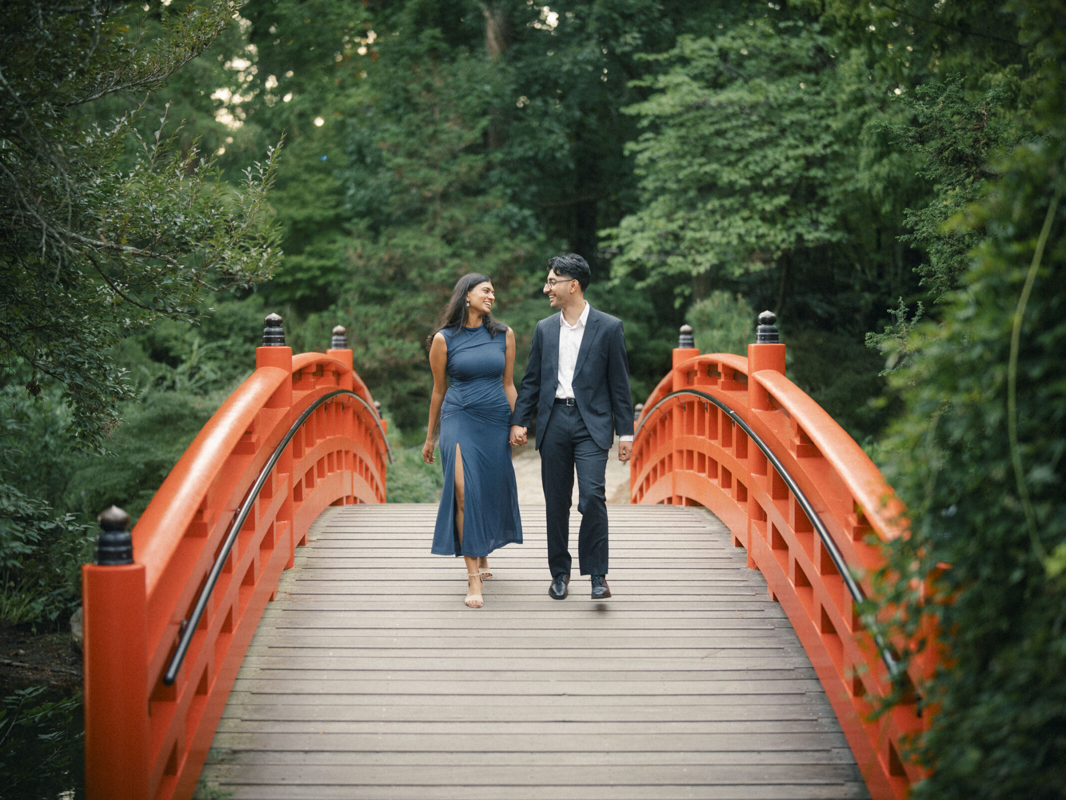 A couple holds hands and smiles at each other while walking across an arched red wooden bridge in Duke Gardens during an engagement photography session. By award-winning Raleigh wedding photographer, Joe Payne.