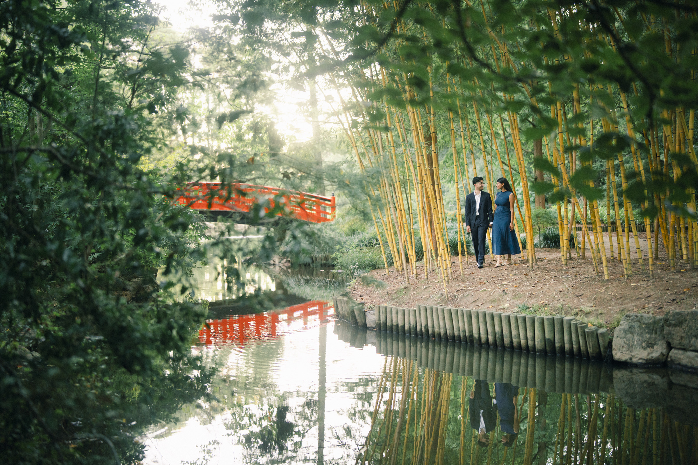 A couple walks through a Duke Gardens bamboo grove beside a pond with a red wooden bridge in the background during their engagement photography session. By award-winning Raleigh wedding photographer, Joe Payne.