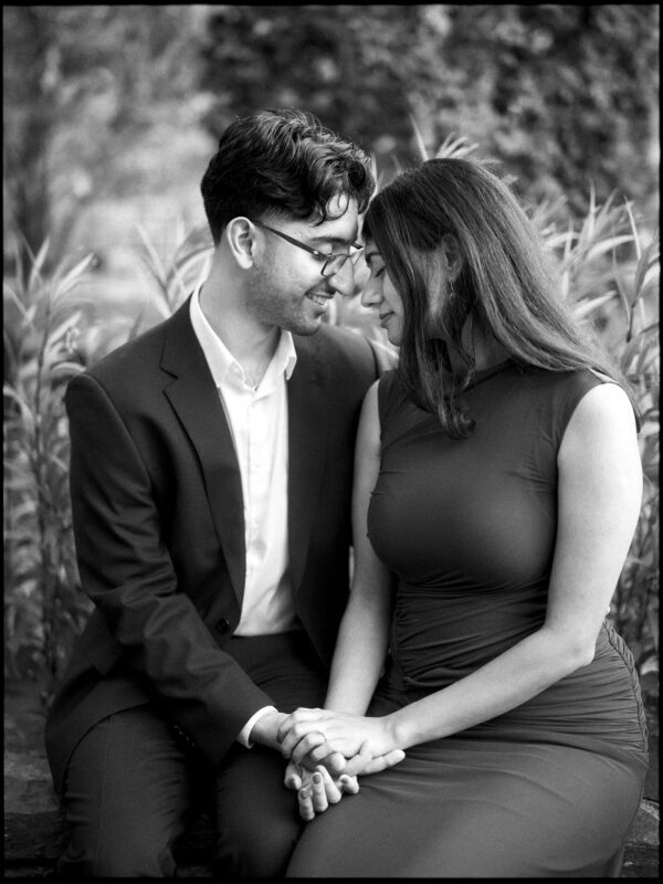 A man in a suit and a woman in a dress sit close together at Duke Gardens, smiling and holding hands during their engagement photography session. By award-winning Raleigh wedding photographer, Joe Payne.
