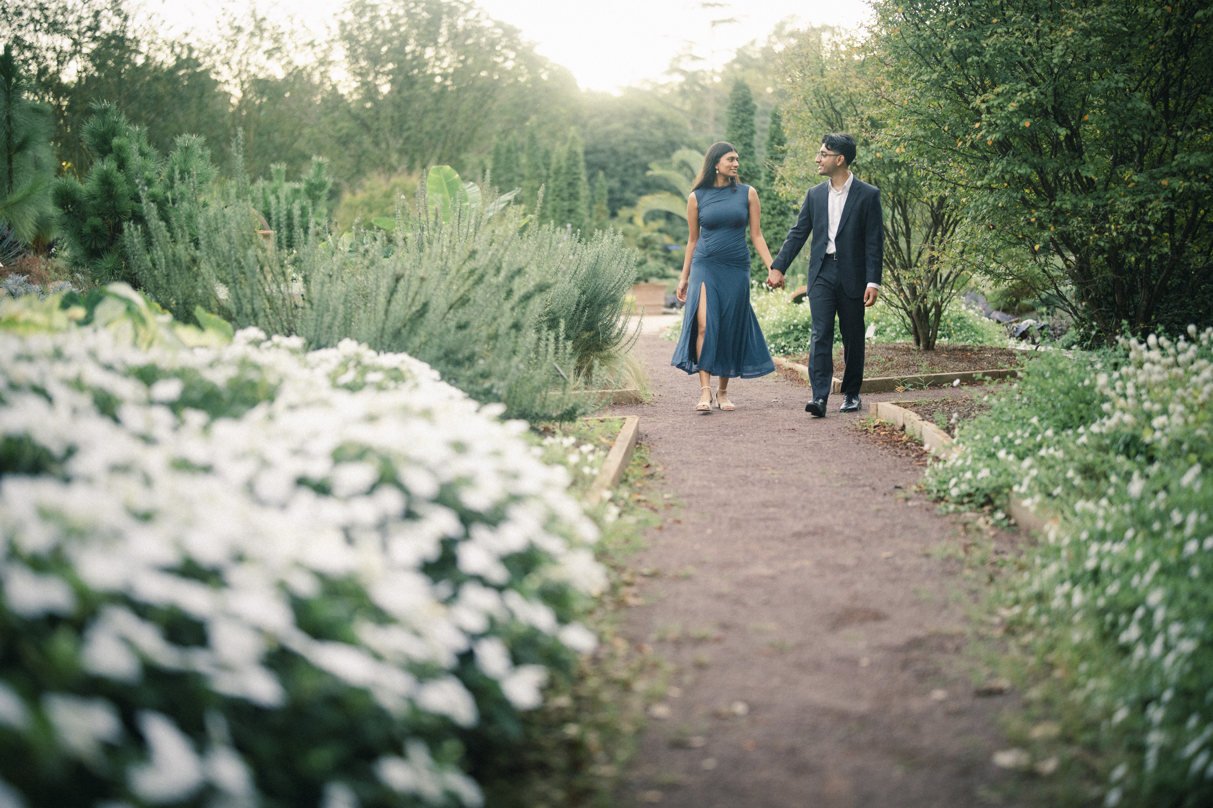 A couple in formal attire holds hands while walking along a gravel garden path during an engagement photography session at Duke Gardens. By award-winning Raleigh wedding photographer, Joe Payne.