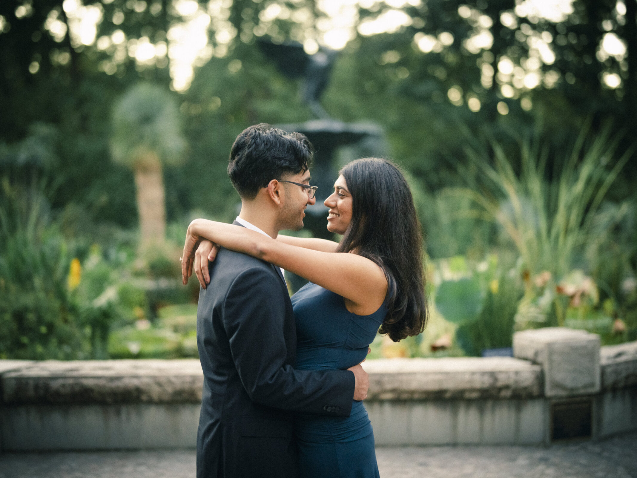 A man in a suit and a woman in a sleeveless dress embrace while looking at each other during a proposal photography session in a park. By award-winning Raleigh wedding photographer, Joe Payne.