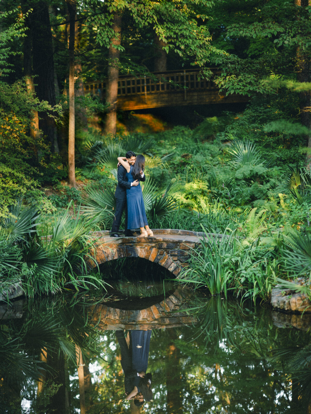 A couple shares a Duke Gardens proposal embrace on a stone footbridge surrounded by lush greenery, with their reflection visible in the water below. By award-winning Raleigh wedding photographer, Joe Payne.