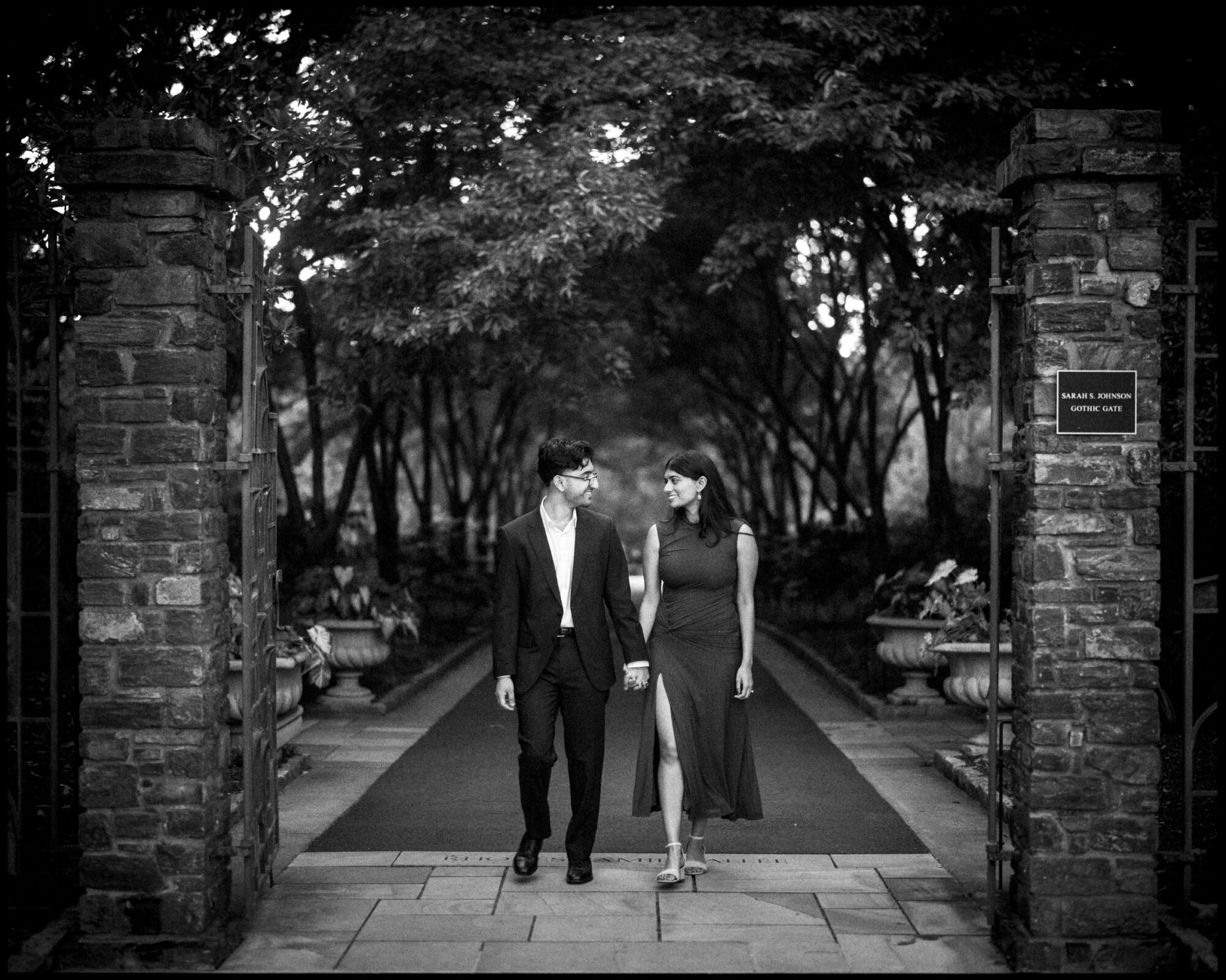 A couple holds hands while walking through a stone gate at Duke Gardens toward the viewer. By award-winning Raleigh wedding photographer, Joe Payne.