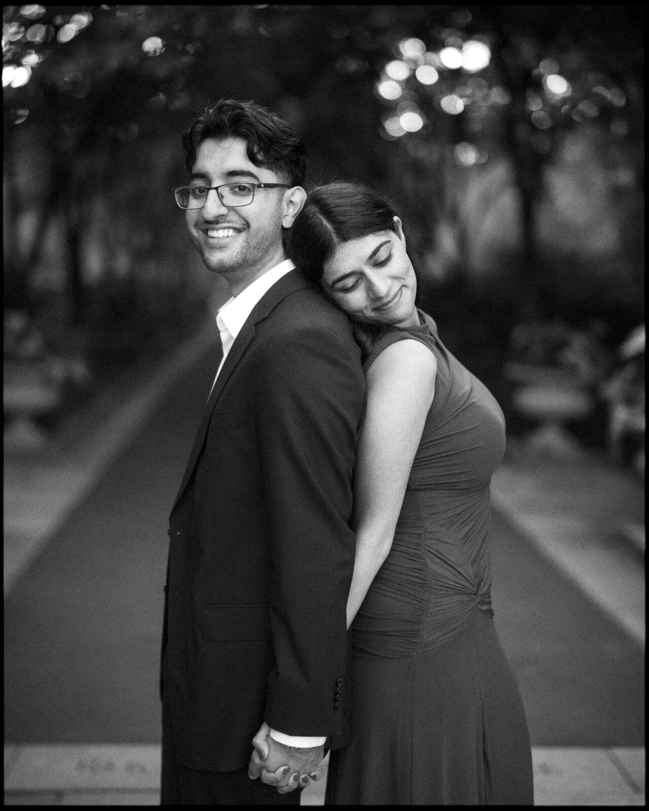 A man in a suit and a woman in a dress stand back-to-back, holding hands during a Duke Gardens engagement photography session. By award-winning Raleigh wedding photographer, Joe Payne.