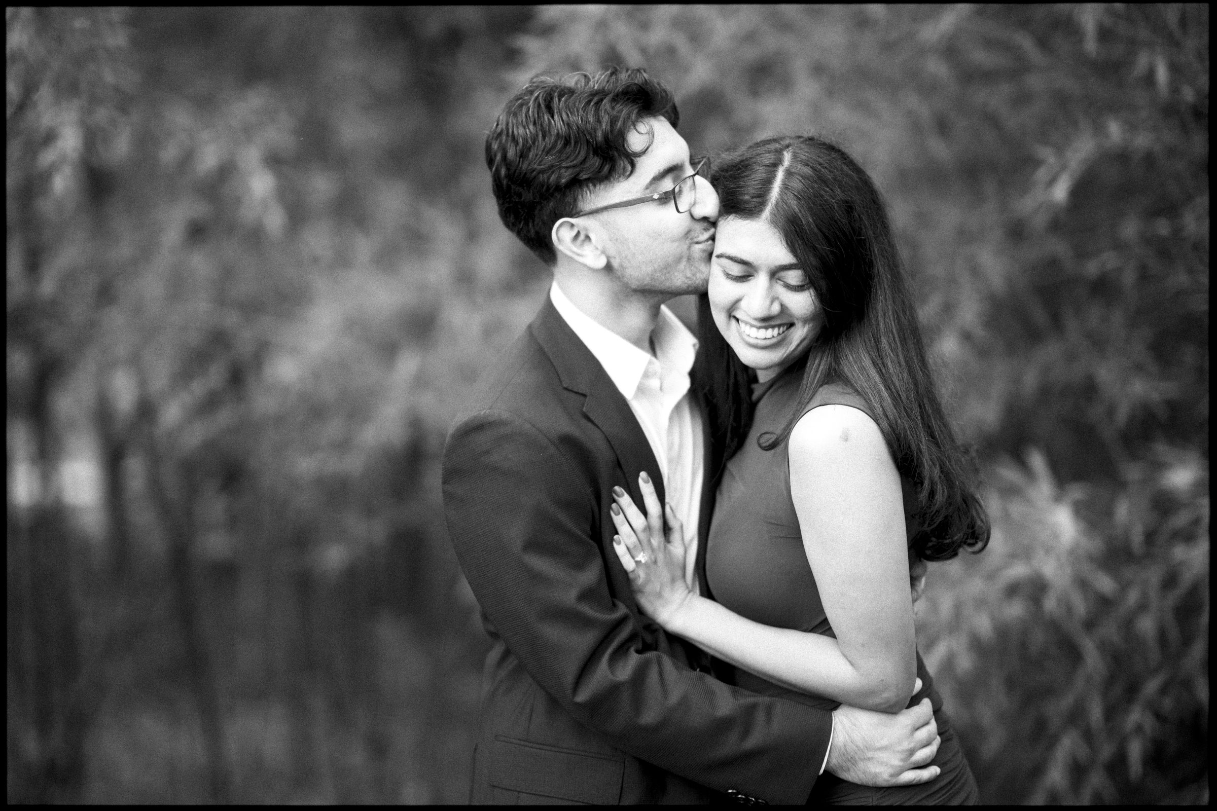 A man in a suit jacket kisses a smiling woman on the cheek during a Duke Gardens proposal as he embraces her. By award-winning Raleigh wedding photographer, Joe Payne.