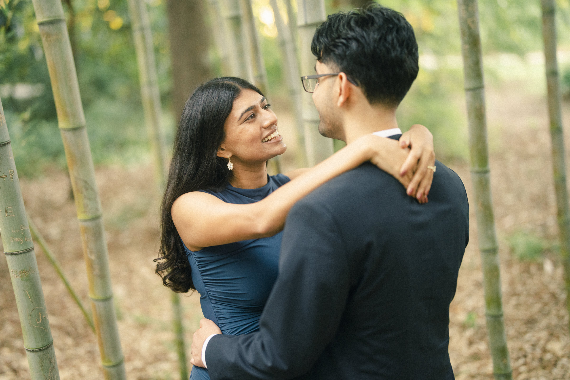A woman in a blue dress gazes affectionately at a man in a dark suit as they embrace in a bamboo grove during an engagement photography session. By award-winning Raleigh wedding photographer, Joe Payne.